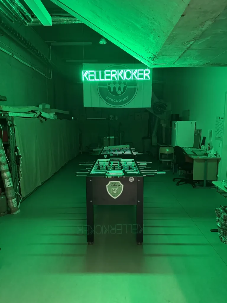 Der Kickerkeller im grünen Licht, über den Tischen das Neonschild „Kellerkicker“ und die Vereinsfahne